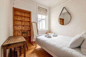 A bedroom with a wooden desk, a chair, a bed with white bedding, a round mirror, and a window with blinds. - Woodburn Terrace (No 8) | St Andrews (St Andrews)