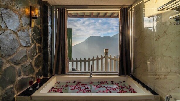 Uma casa de banho com parede de pedra, uma banheira com pétalas de rosa, vista para montanhas e uma vedação de madeira.