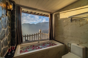 A bathroom with a bathtub, a window with a view of mountains, and a stone wall. - Highlanders Cafe Stay (Mussoorie)