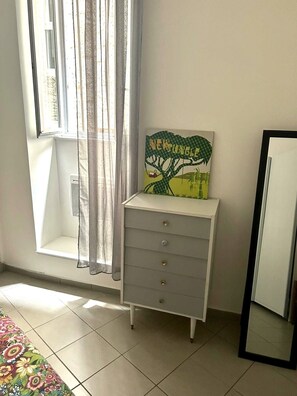 Una habitación con ventana, un aparador blanco, un espejo y un cuadro de una escena selvática.