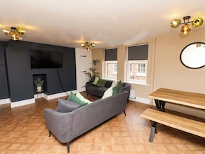Ein Wohnzimmer mit grauem Sofa, einer Holzbank, einem an der Wand befestigten Fernseher und einem Fenster mit Jalousien.