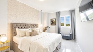 Ein Schlafzimmer mit einem großen Bett, einem Nachttisch, einem Fenster mit Vorhängen und einem an der Wand montierten Fernseher.