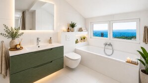 Ein modernes Badezimmer mit großem Spiegel, einer Badewanne, einer Toilette und einem Fenster mit Meerblick.