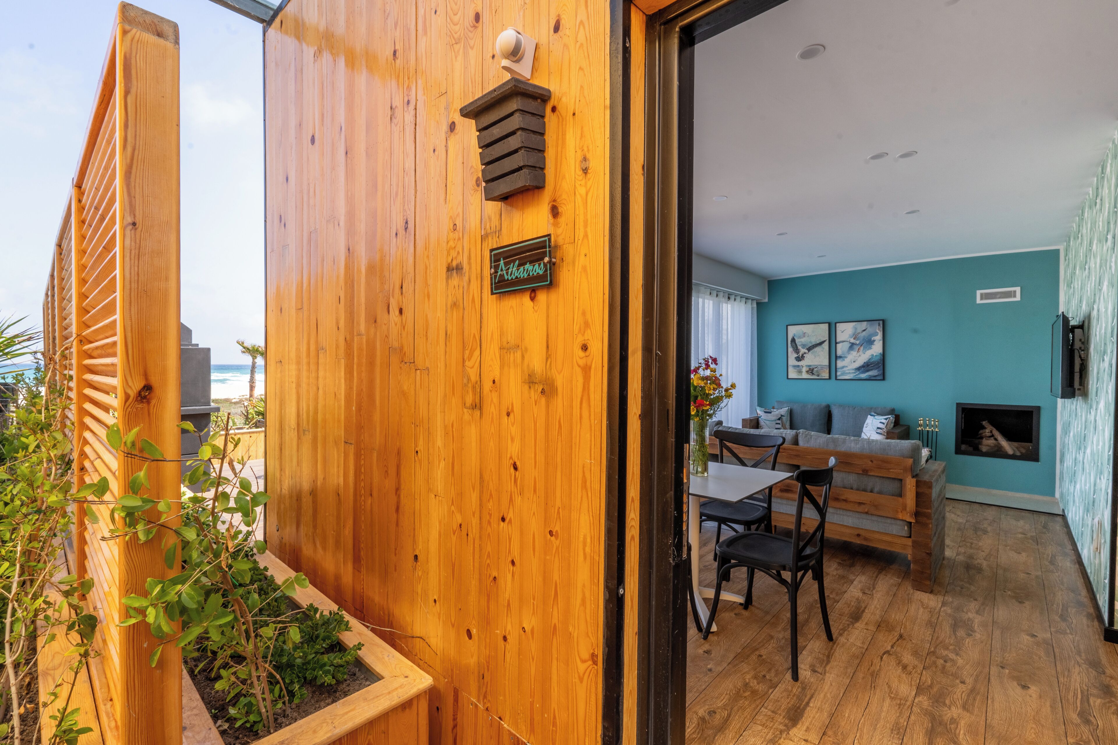 Een houten buitenmuur met een deur die leidt naar een kamer met een blauwe wand, een open haard en een eethoek met een tafel en stoelen.