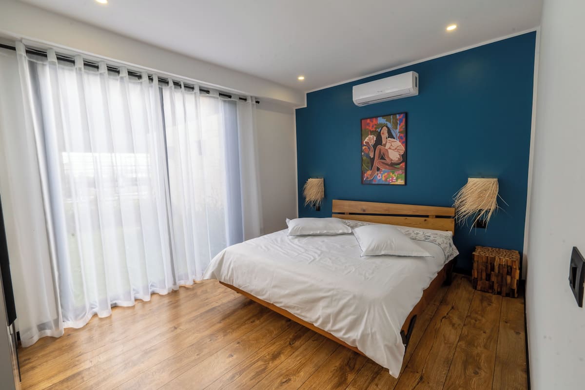 Un dormitorio con piso de madera, una cama con ropa de cama blanca, una pared azul y un equipo de aire acondicionado.
