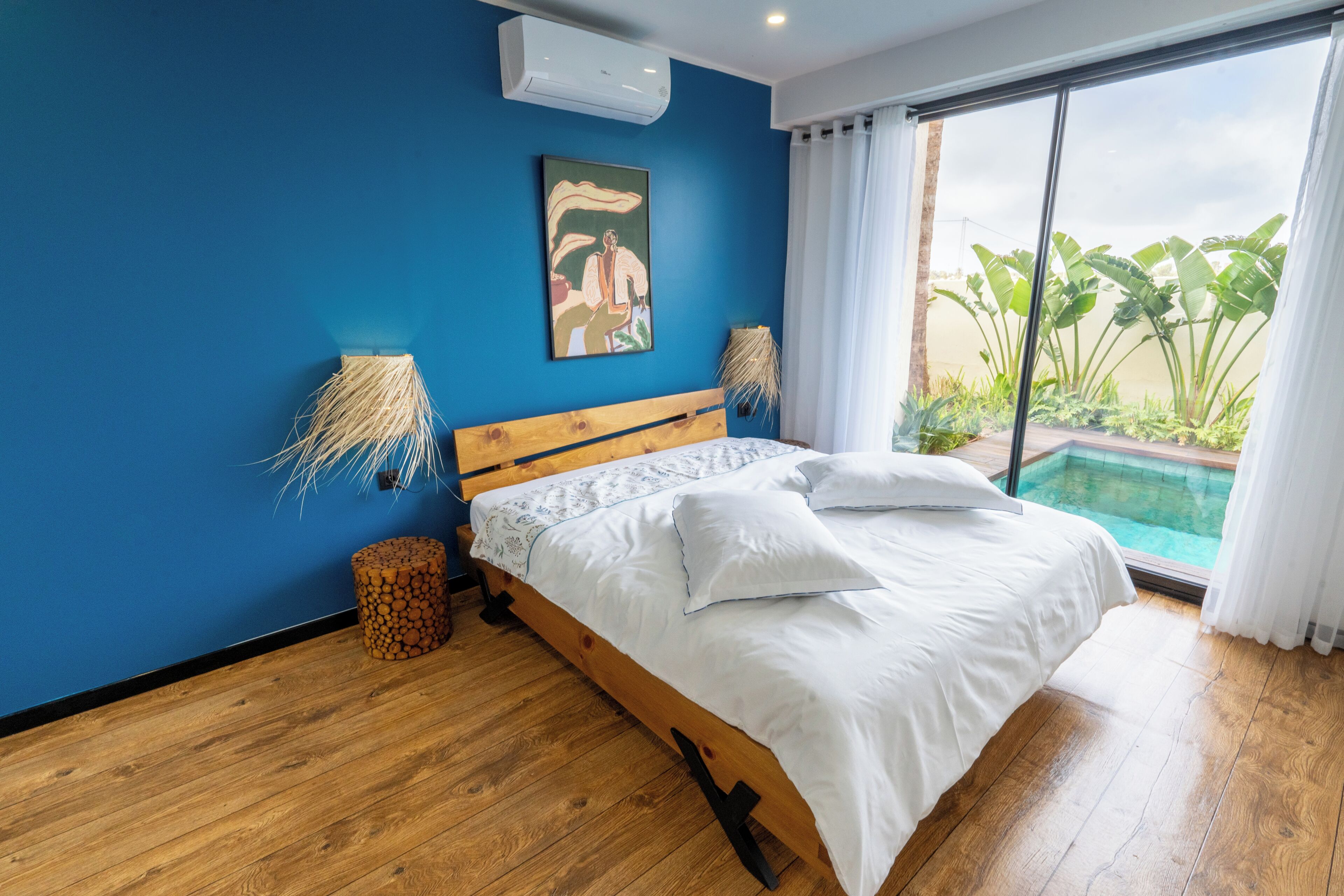 Un dormitorio con cama de madera, pared azul, un cuadro, una ventana con vistas a plantas y una piscina.