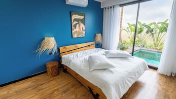 Un dormitorio con cama de madera, pared azul, un cuadro, una ventana con vistas a plantas y una piscina.