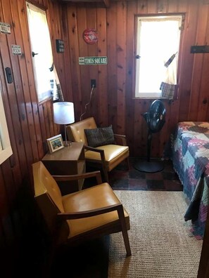 Een kamer met houten wanden, een bureau met lamp, twee stoelen, een bed met quilt, een ventilator en een raam met uitzicht op een gebouw.