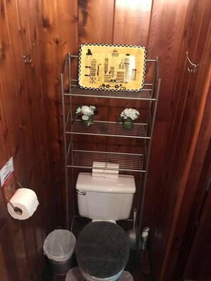 Een kleine badkamer met toilet, een metalen plankenunit, een prullenbak en een rol toiletpapier.