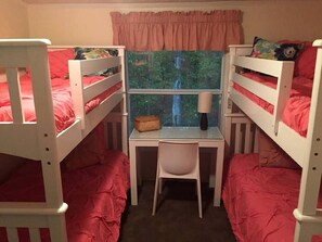 Een slaapkamer met stapelbed, bureau en stoel, een raam met uitzicht op bomen en een roze gordijn.
