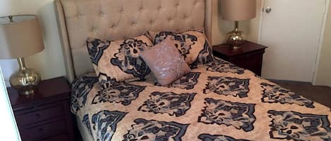 Un dormitorio con cabecera capitonada, colcha estampada, dos lámparas de noche y una mesita de noche.