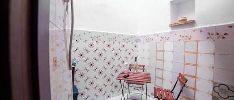 A small, enclosed patio area with a table and chairs, tiled walls with a floral pattern, and a shelf with a bowl.