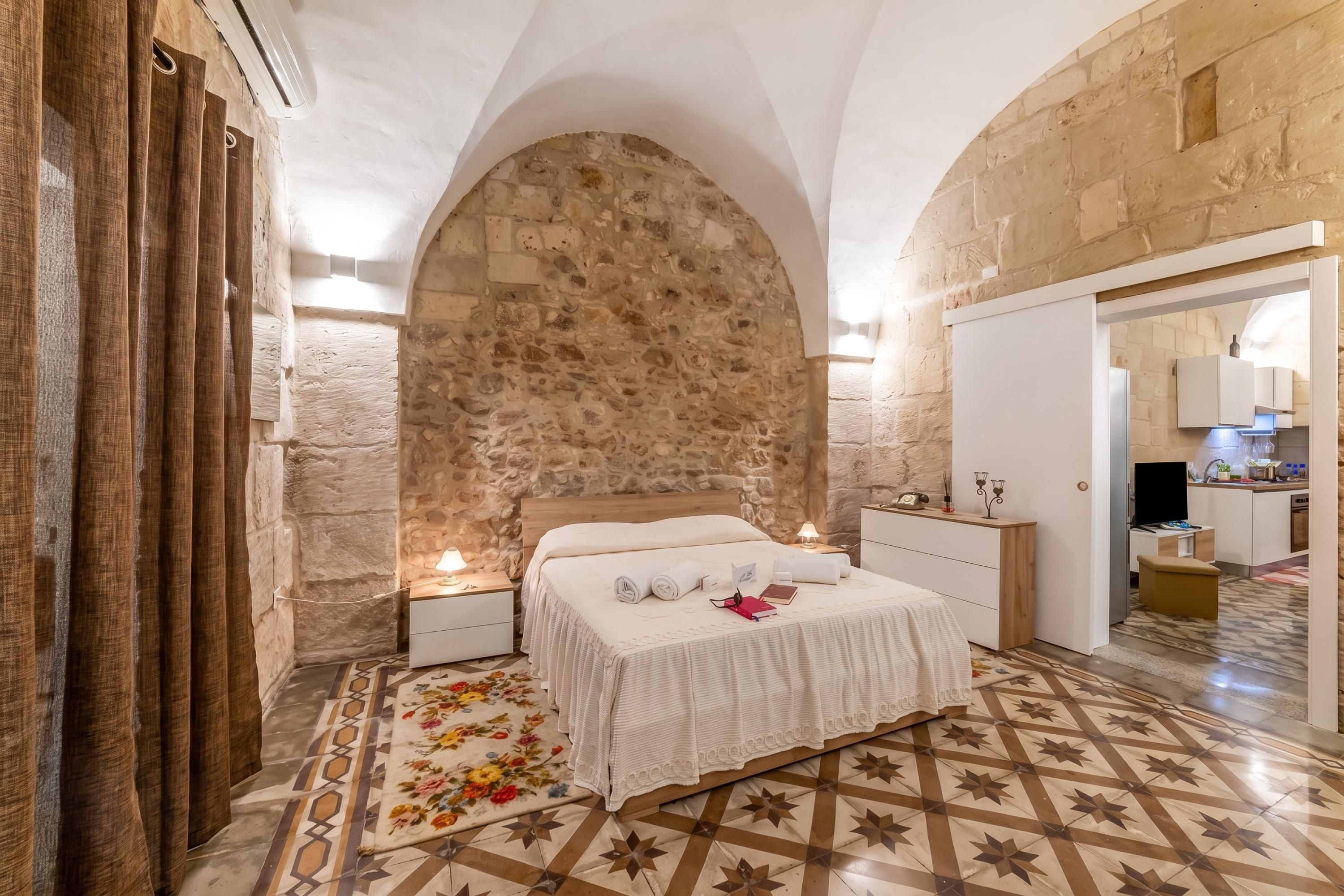 Une chambre avec un mur en pierre, un lit recouvert d’un tapis à motifs floraux, et une cuisine visible à travers une porte ouverte.