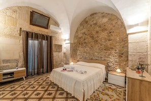 Ein Zimmer mit Steinwand, einem Bett mit weißer Bettdecke, einem Nachttisch mit Lampe und einer Holzkommode.