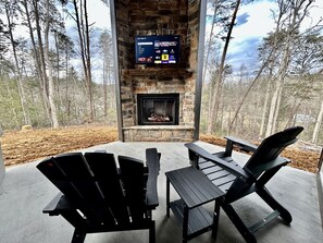 A stone fireplace with a TV screen above it, surrounded by Adirondack chairs on a concrete deck with a forest backdrop. - Romantic Cabin in Shady Grove Area of the Smoky Mountains: A Perfect One-Level Retreat for Couples (Sevierville)