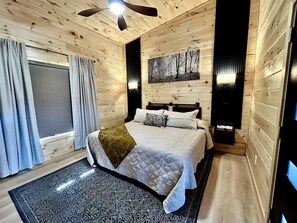 A bedroom with a bed, a ceiling fan, a window with curtains, and a painting on the wall. - Romantic Cabin in Shady Grove Area of the Smoky Mountains: A Perfect One-Level Retreat for Couples (Sevierville)