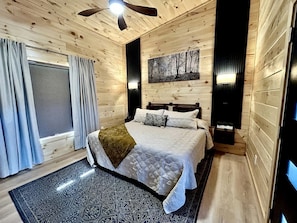 A bedroom with a bed, a ceiling fan, a window with curtains, and a painting on the wall. - Romantic Cabin in Shady Grove Area of the Smoky Mountains: A Perfect One-Level Retreat for Couples (Sevierville)