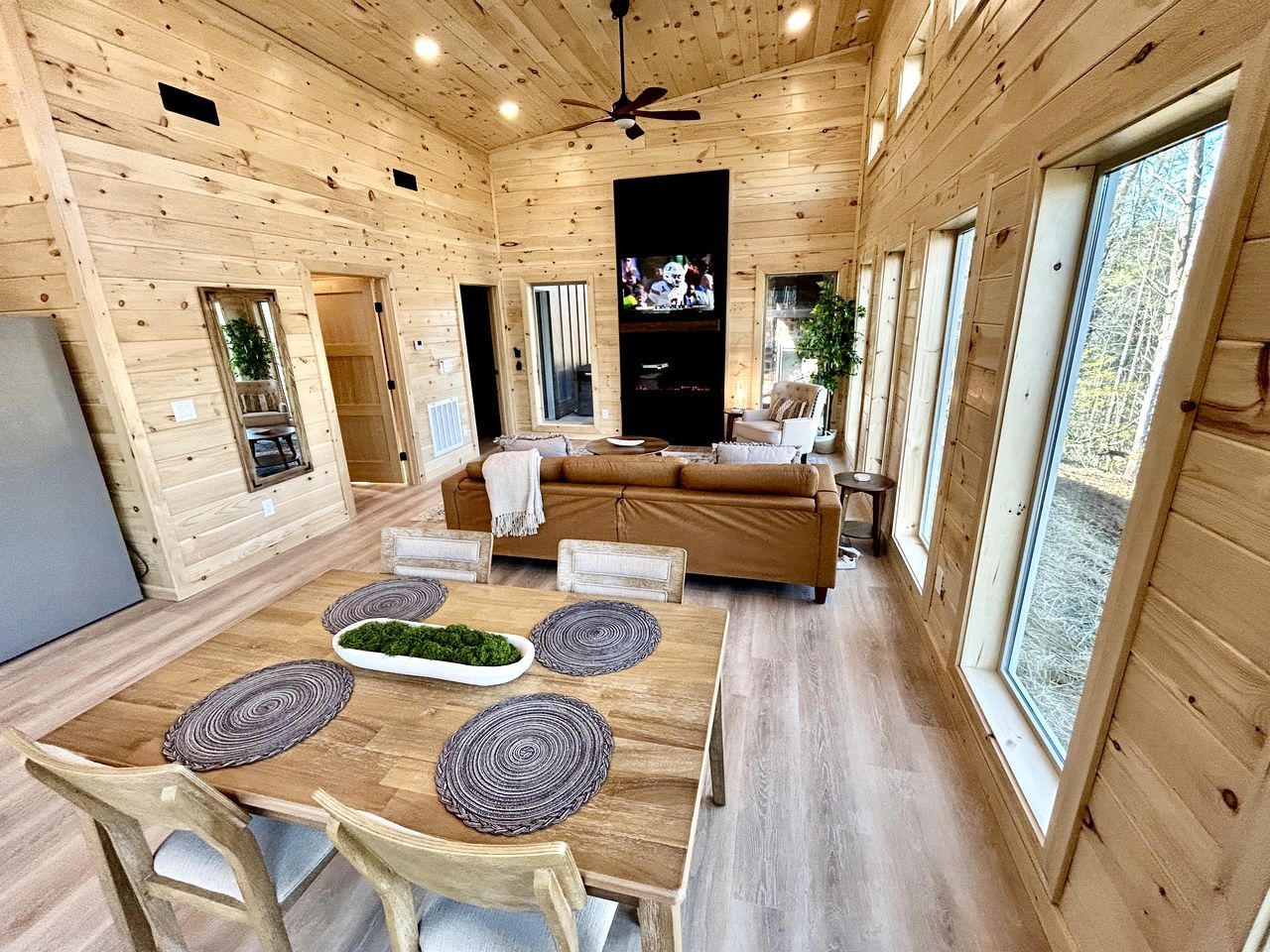 Interior d'una cabana de fusta amb menjador, sofà, televisió i finestres grans.