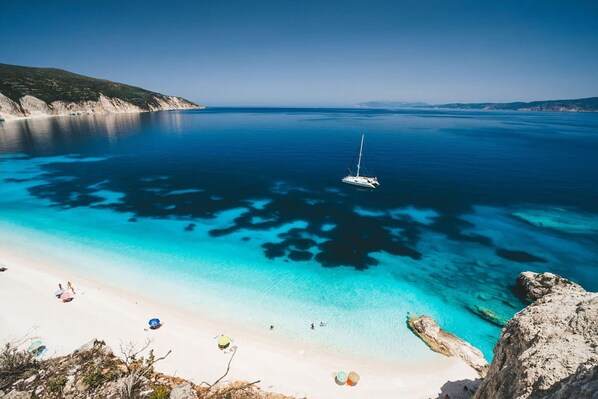 A coastal beach with clear turquoise waters, a sailboat, and rocky outcrops.