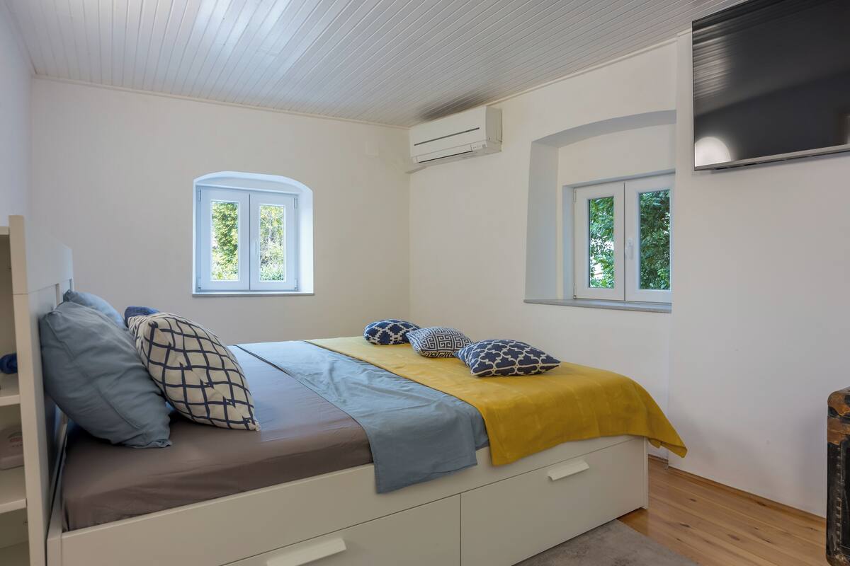 A bedroom with a bed, two windows, an air conditioning unit, and a television on the wall.