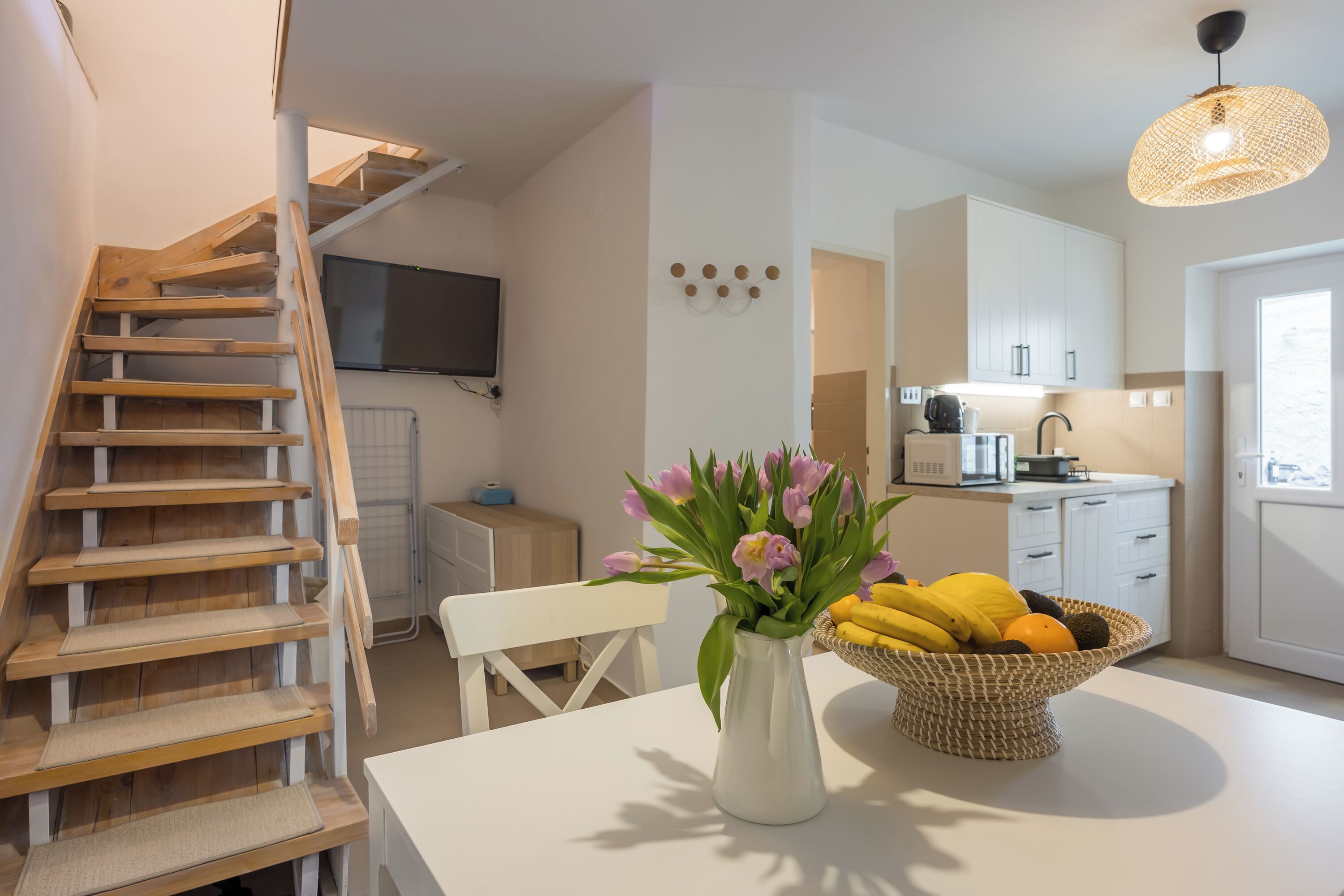 A modern kitchen with white cabinets, a wooden staircase, a flat-screen TV, and a vase with flowers on a table.