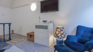 Ein Zimmer mit einem wandmontierten Flachbildfernseher, einer weißen Kommode mit Schubladen, einem blauen Sessel, einem gemusterten Teppich und einer Lampe mit floralem Dekor.