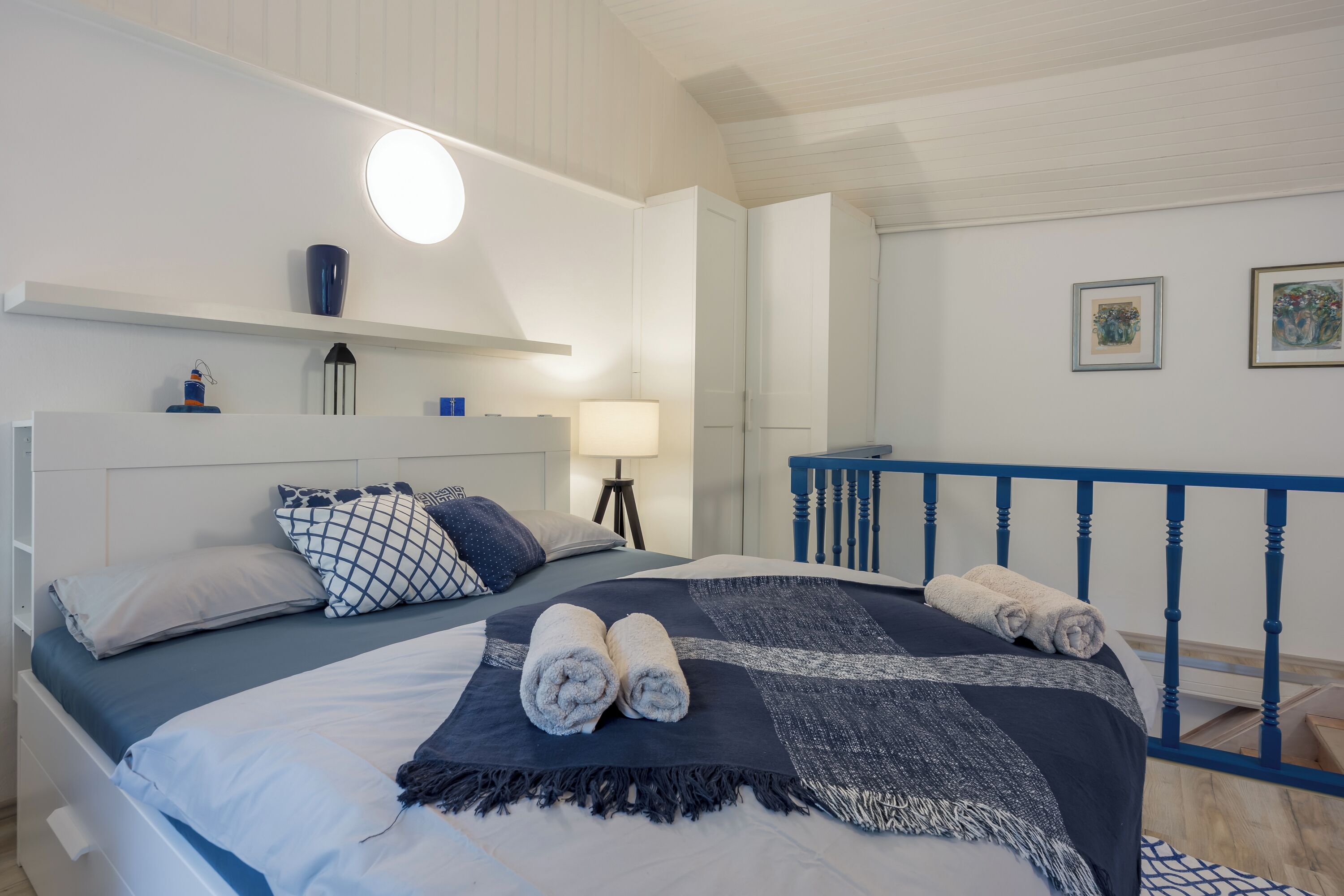 A neatly made bed with rolled towels, a bedside lamp, and a wall-mounted shelf with decorative items.