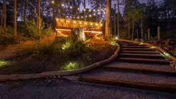 A wooden deck with string lights, surrounded by trees and a gravel path leading to it.
