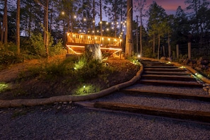 Una terraza de madera con luces colgantes, rodeada de árboles y un sendero de grava que conduce a ella.