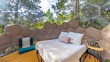 A bedroom with a bed, a chair, a small table, and a view of trees through a geometric window structure.