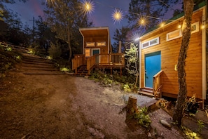 Una cabaña con un muelle de madera y una puerta azul, rodeada de árboles y un sendero de tierra.
