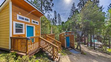 A wooden cabin with a deck, a blue door, and a small shed in a forested area.