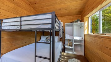 Een slaapkamer met een stapelbed, houten plafond, raam met uitzicht op bomen en een witte garderobe met opgevouwen kleding.