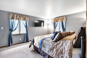 A bedroom with a large bed, two windows with curtains, a flat-screen TV, and a carpeted floor.