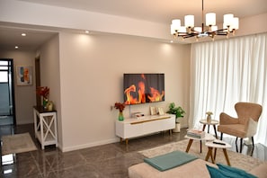 A modern living room with a flat-screen TV, a chandelier, a sofa, a coffee table, and a side table with a remote control. - Sky homes 3bedroom with a park view (Syokimau)