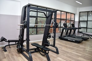 Un gimnasio con equipo de levantamiento de pesas, cintas de correr y ventanas amplias.