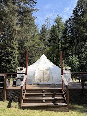 A white tent with a wooden staircase leading up to it, surrounded by a wooden deck and dense forest.