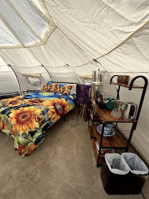 A tented room with a bed, a shelf with kitchen items, and a trash bin.