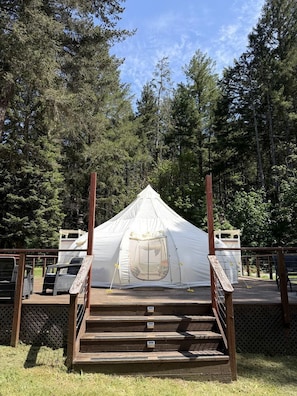A white tent with a wooden staircase leading up to it, surrounded by a wooden deck and dense forest.