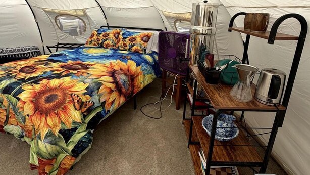 Una habitación con carpa, una cama, un estante con utensilios de cocina y un cubo de basura.