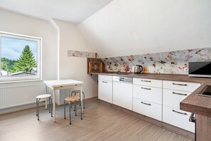 A compact kitchen with a white countertop, wooden stools, a dishwasher, and a window with a view of trees. - Apartment Herian with Shared Garden on the Edge of Müritz National Park (Schloen-Dratow)