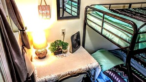 A bunk bed with a green and black patterned bedspread, a small table with a lamp, a fan, and a framed mirror.