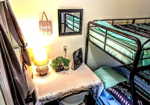 A bunk bed with a green and black patterned bedspread, a small table with a lamp, a fan, and a framed mirror.