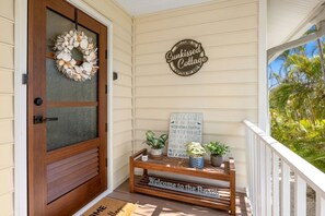 Una puerta de madera con un panel de vidrio esmerilado, un letrero de bienvenida, un pequeño banco de madera con plantas en macetas y un felpudo que dice 'Welcome to the Beach'.