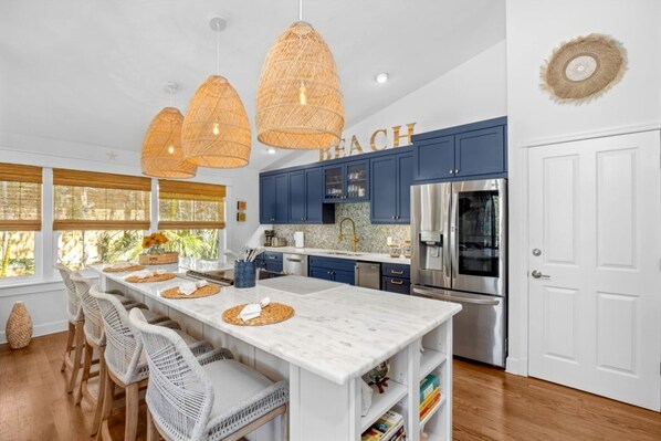 Una cocina moderna con una isla central, armarios azules, electrodomésticos de acero inoxidable, lámparas colgantes de mimbre y vistas al exterior.