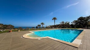 Una piscina de borde curvo, rodeada por una terraza con sillones de descanso y plantas en macetas, con vistas a un paisaje costero.