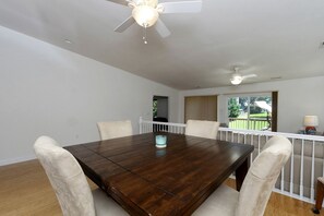 Comedor con mesa de madera oscura, sillas blancas acolchadas, ventiladores de techo y vista a un patio trasero a través de una ventana.