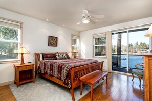 A bedroom with a bed, bedside tables, a bench, a ceiling fan, and large windows with a view of a body of water.