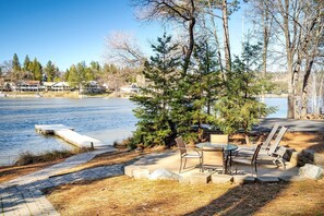 A lakeside patio with a dock, outdoor seating, and a view of houses across the water.