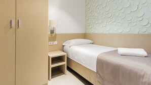 Una cama individual con un almohadón blanco y una manta color beige, una mesita de noche con una lámpara, y una pared con un diseño circular.